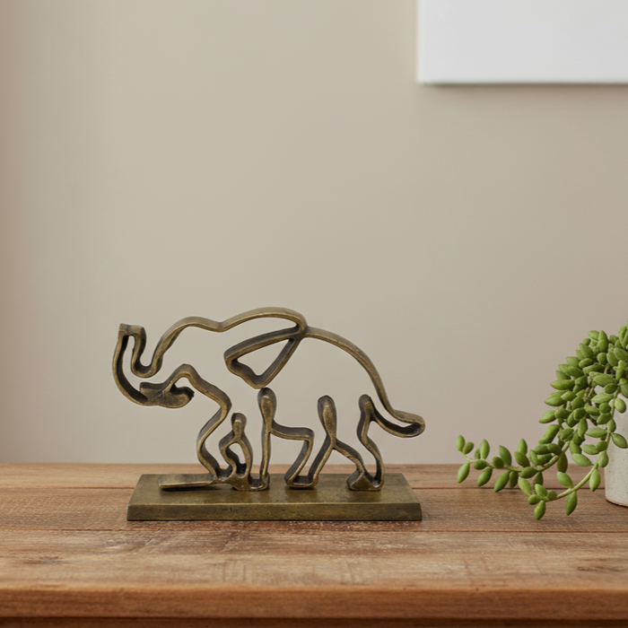 Dekorative Elefantenfigur aus Metall mit Patina auf Sockel, minimalistisch inszeniert auf Holztisch neben grüner Pflanze vor hellem Hintergrund.