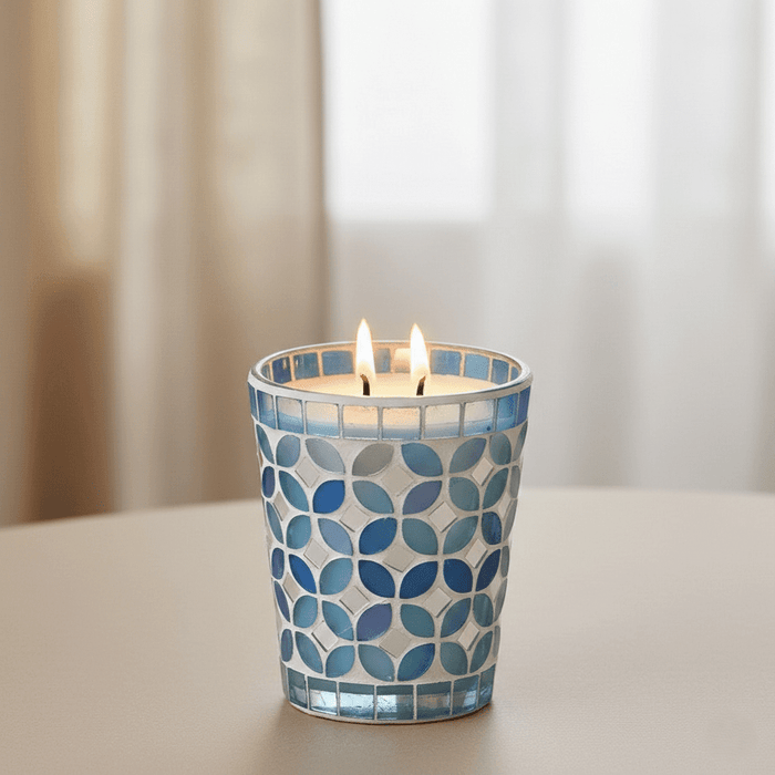 Duftkerze im blauen Mosaikglas mit zwei Flammen, dekorativ auf hellem Tisch vor weissem Vorhang.