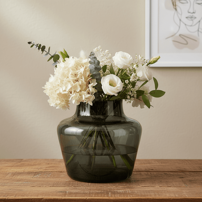 Mundgeblasene Glasvase in Rauchgrau mit weissem Blumenstrauss aus Hortensien und Rosen, dekorativ auf einem Holztisch vor hellem Hintergrund.