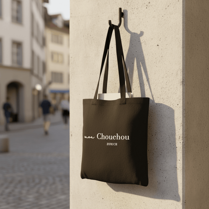Schwarze Tote Bag mit Mon Chouchou Zürich Logo in Weiss, stilvoll an Betonwand aufgehängt in urbaner Zürcher Umgebung.